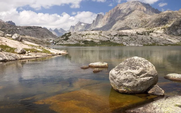HD desktop wallpaper of a clear mountain stream flowing through rocky terrain with large boulders, surrounded by rugged peaks under a partly cloudy sky.