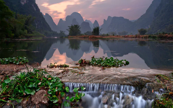 Serene landscape of the Nanling Mountains in Guangxi Zhuang, China, featuring calm water reflections, lush greenery, and misty mountain peaks at sunset.
