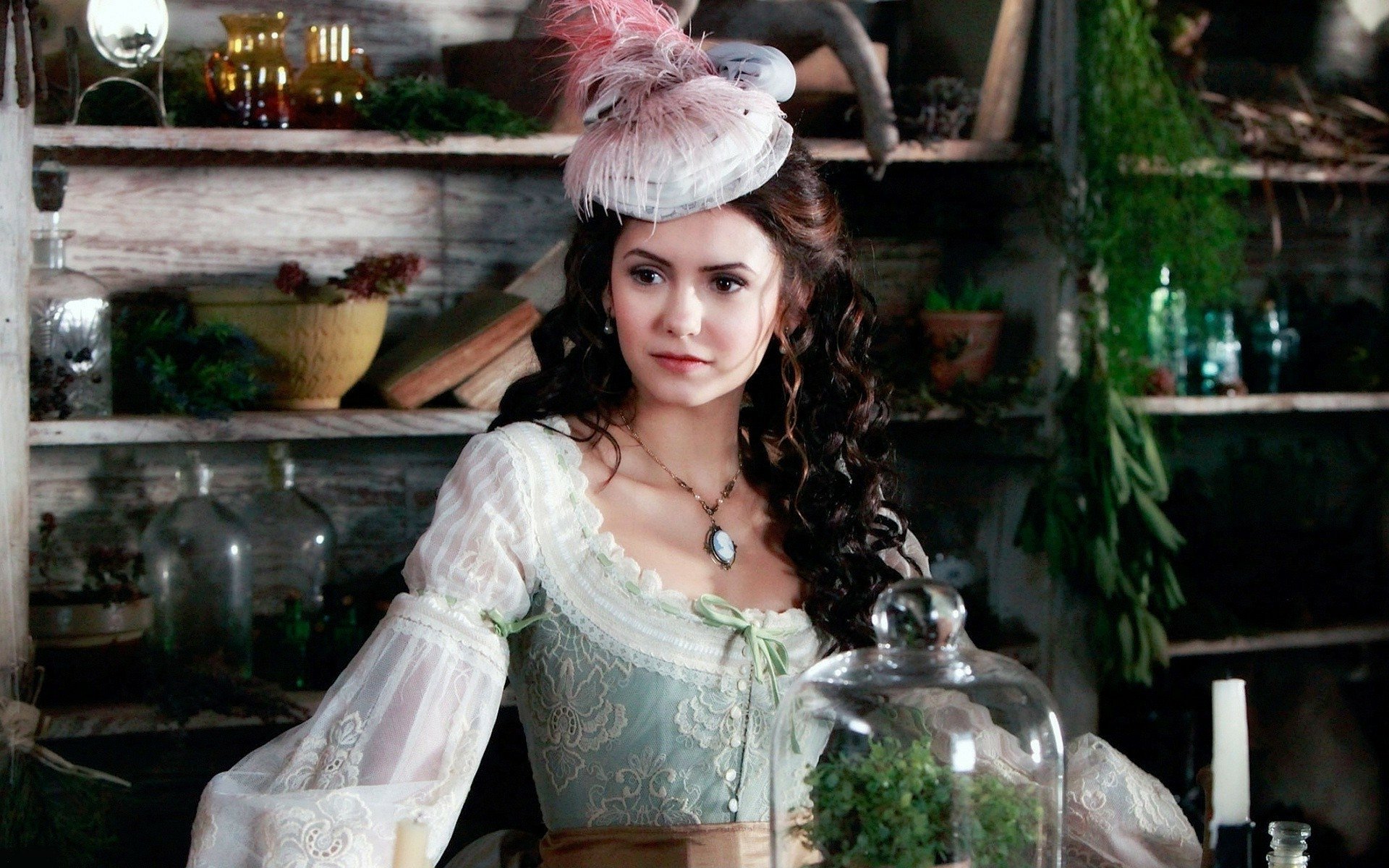 HD desktop wallpaper featuring celebrity Nina Dobrev dressed in an elegant, vintage-style dress with a feathered hat, set in a rustic, apothecary-like background.
