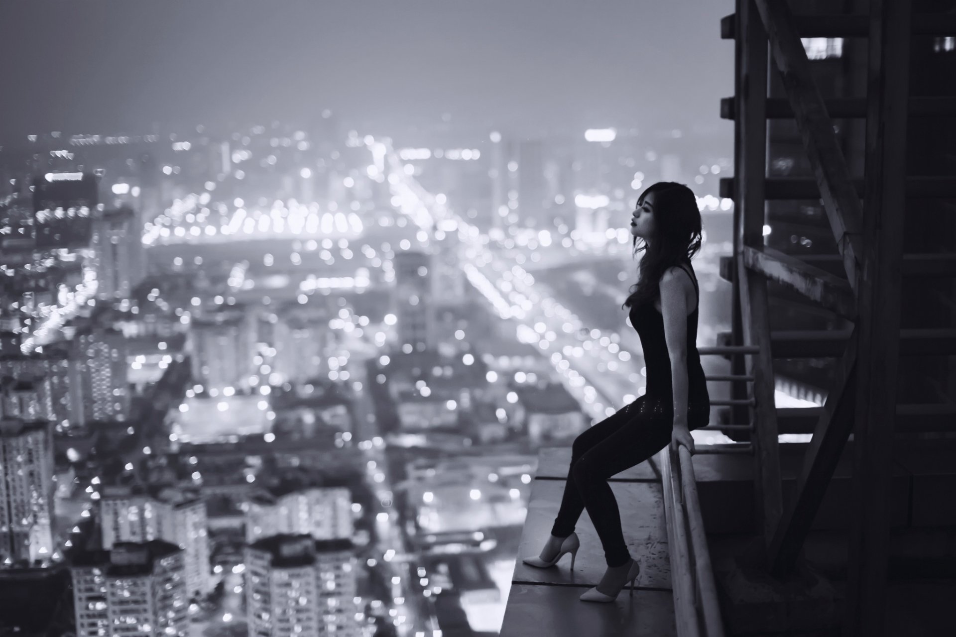 HD desktop wallpaper featuring an Asian woman sitting on a rooftop edge, overlooking a brightly lit cityscape at night.