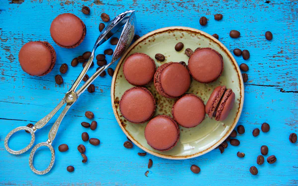 5K Ultra HD PC desktop wallpaper of food: chocolate macarons on a plate, one halved, with scattered coffee beans and silver sugar tongs on a bright turquoise wooden background.