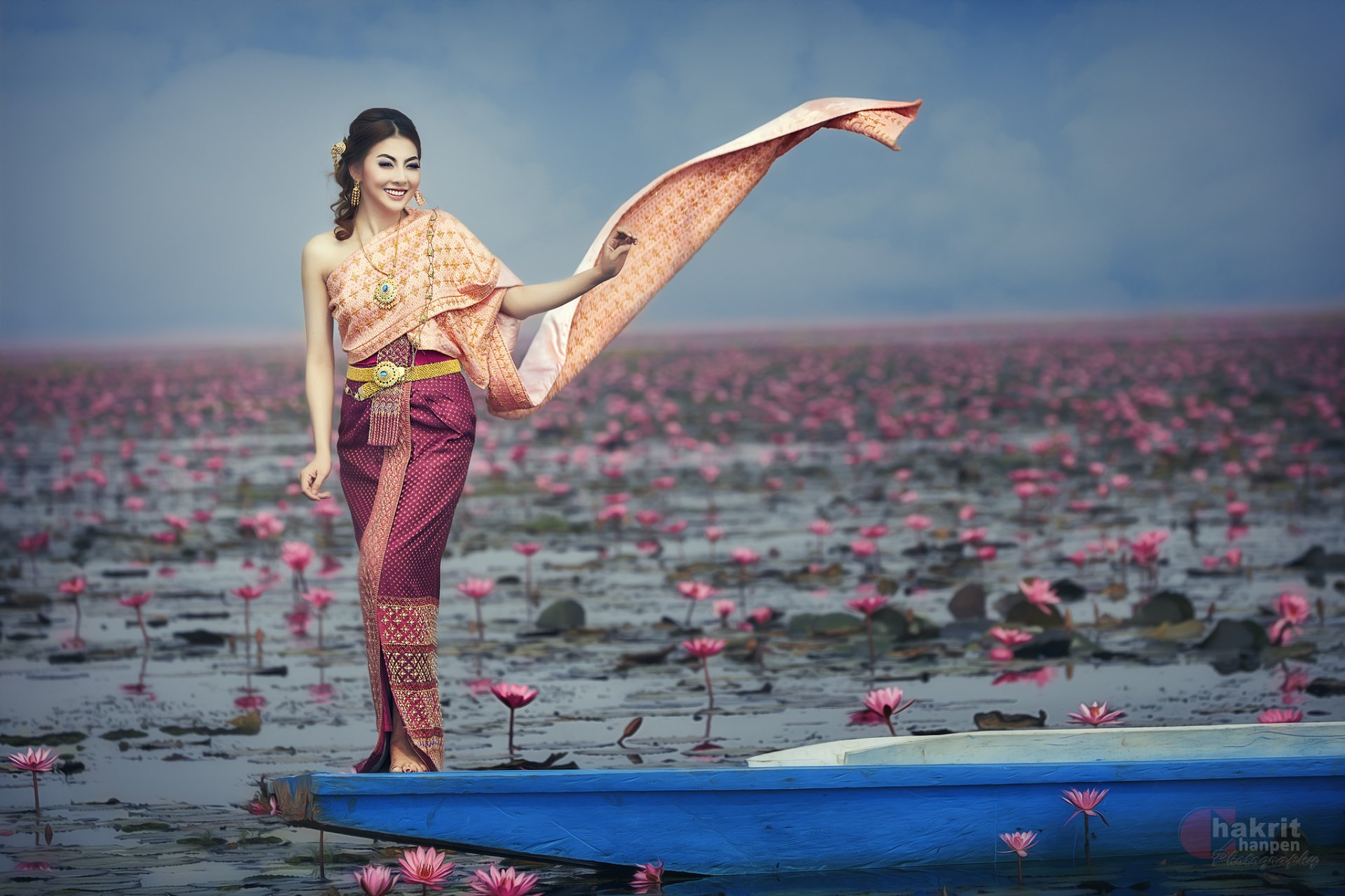 A woman in traditional Thai national dress stands gracefully on a boat surrounded by blooming lotus flowers, captured in vibrant HD photography against a serene backdrop.