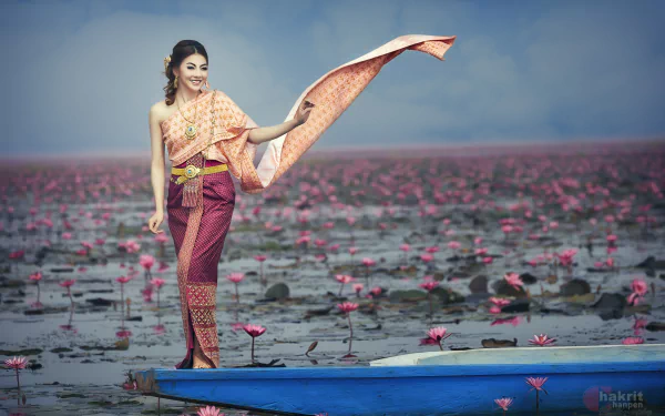 A woman in traditional Thai national dress stands gracefully on a boat surrounded by blooming lotus flowers, captured in vibrant HD photography against a serene backdrop.