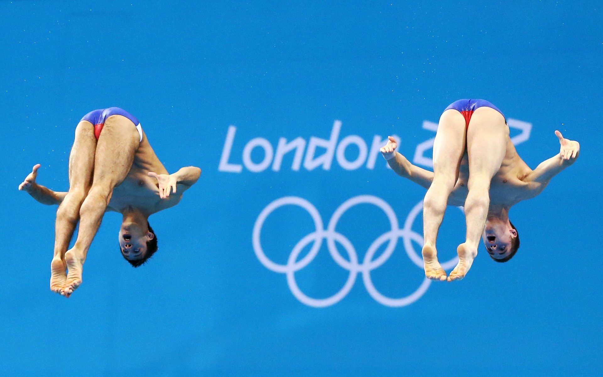 2K Quad HD PC desktop wallpaper of two synchronized divers frozen midair against a blue backdrop with London 2012 text and Olympic rings — dynamic diving sports action.
