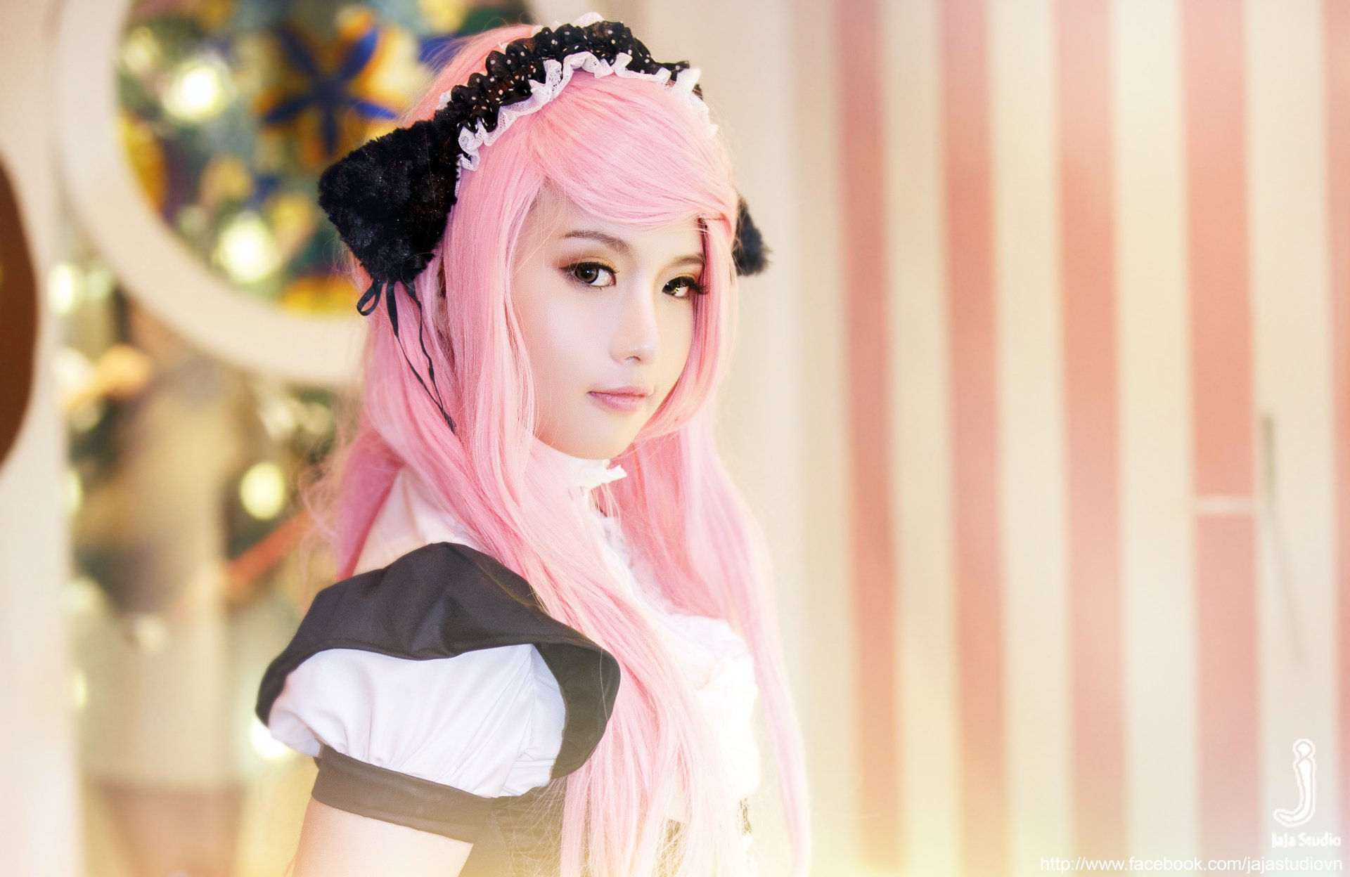 A Vietnamese woman with pink hair in a cosplay outfit poses against a softly lit, striped background, capturing a whimsical and enchanting atmosphere.