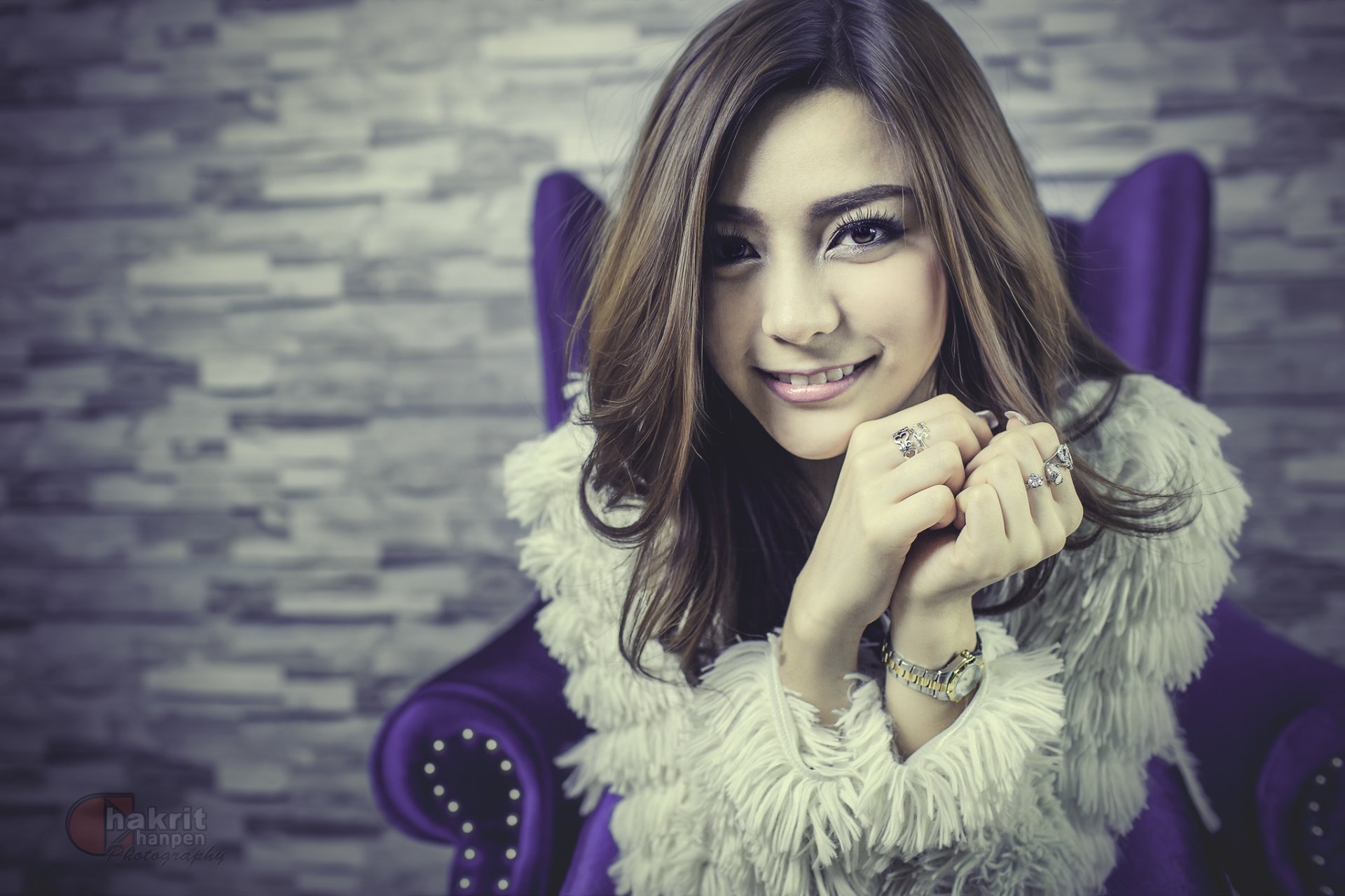 HD desktop wallpaper featuring a smiling Thai Asian woman model in a white fur jacket, seated on a purple chair against a stone wall background.