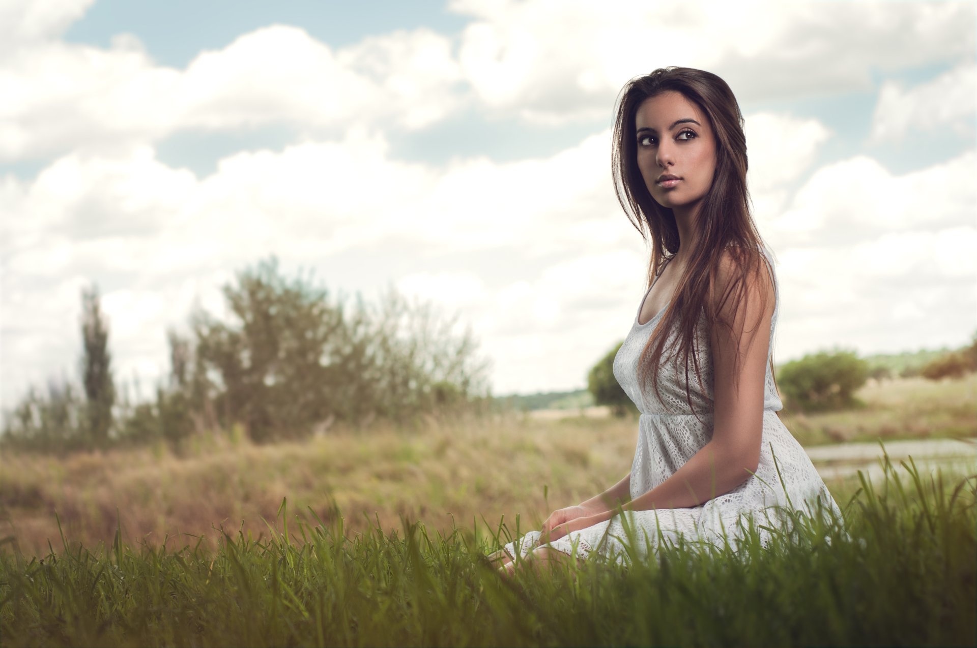 A serene Asian model in a flowing white dress sits gracefully in a lush green field, framed by a bright sky and soft clouds, creating a captivating HD desktop wallpaper background.