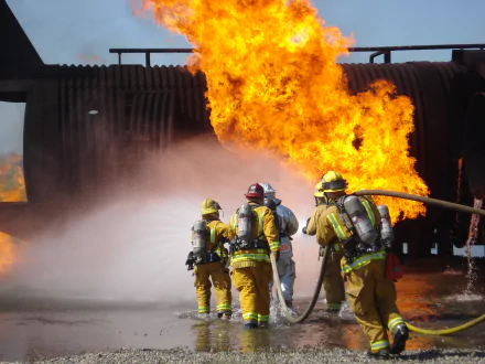 A group of firefighters in yellow gear battles a large fire, with flames and smoke rising dramatically in the background. This intense scene makes for an engaging HD PC desktop wallpaper.