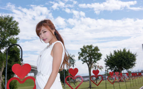 Taiwanese model Xiao Xi stands against a bright sky in a white dress, featured in a 4K Ultra HD desktop wallpaper with heart-shaped decorations lining the park fence.