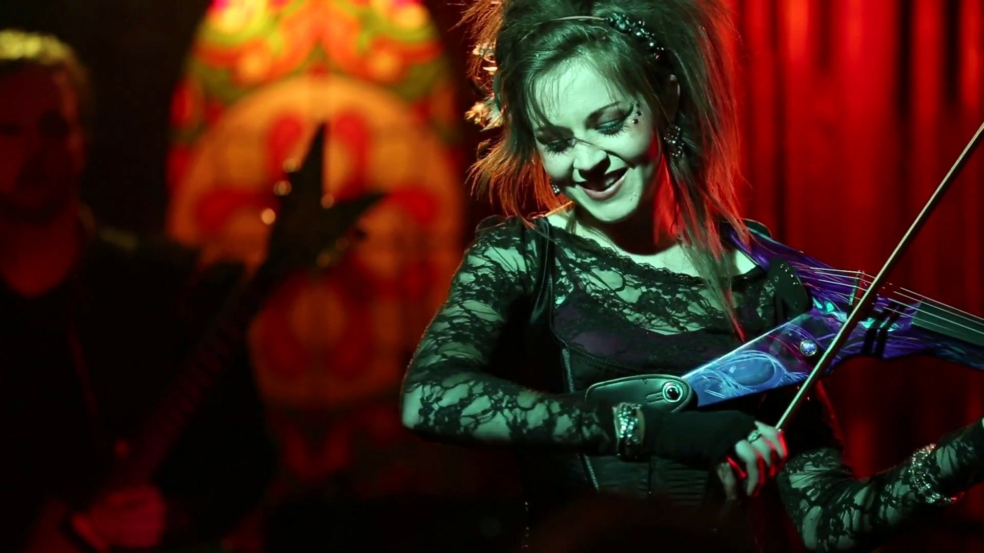 HD PC desktop wallpaper featuring Lindsey Stirling playing the violin on stage, bathed in warm red and orange lighting with a vibrant background.