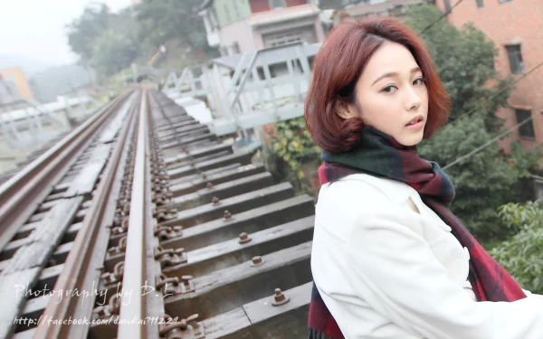 A Taiwanese model stands by a railroad, gazing intently at the camera. The scene captures a serene, misty atmosphere, highlighting her stylish outfit and the train tracks.