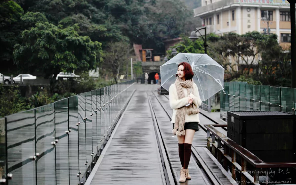 A Taiwanese woman holding an umbrella walks along a rainy pathway, blending into the lush scenery. This HD desktop wallpaper features a model, Lín Yǔ, capturing a serene moment in the rain.