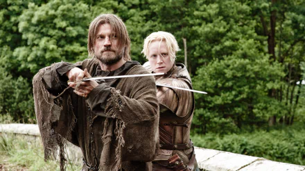 Gwendoline Christie as Brienne of Tarth and Nikolaj Coster-Waldau as Jaime Lannister stand back-to-back, armed with swords, in a scene from *Game of Thrones*, surrounded by lush greenery.