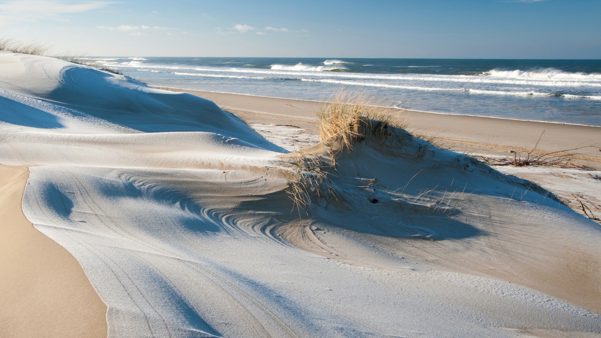 HD PC desktop wallpaper and background: serene beach nature scene with wind-swept sand dunes, grassy tufts, gentle surf and a clear blue sky.