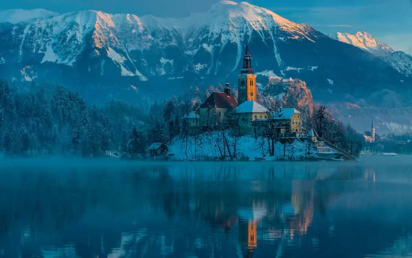 A serene sunrise over Lake Bled reflects the stunning Julian Alps, showcasing the Assumption of Mary Church amidst a wintry landscape in Slovenia.