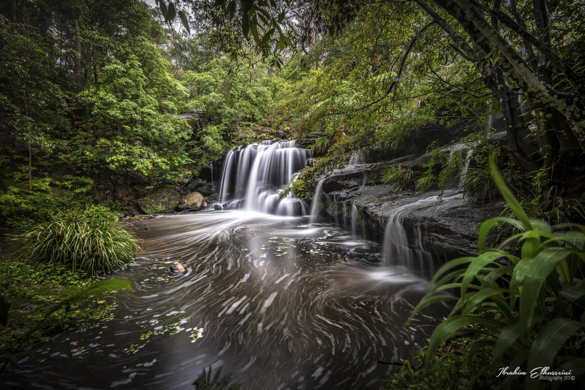 5K Ultra HD PC desktop wallpaper: tranquil Australian forest creek with cascading waterfall, mossy rocks and swirling water beneath a lush green canopy.