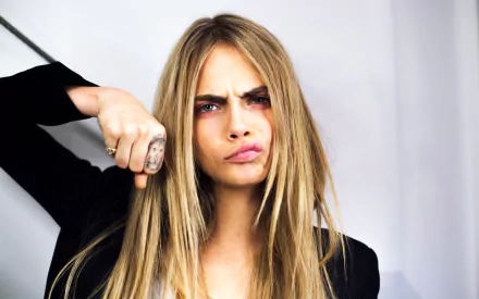 HD desktop wallpaper of English actress and model with blue eyes, Cara Delevingne, displaying a playful expression and long blonde hair against a neutral background.