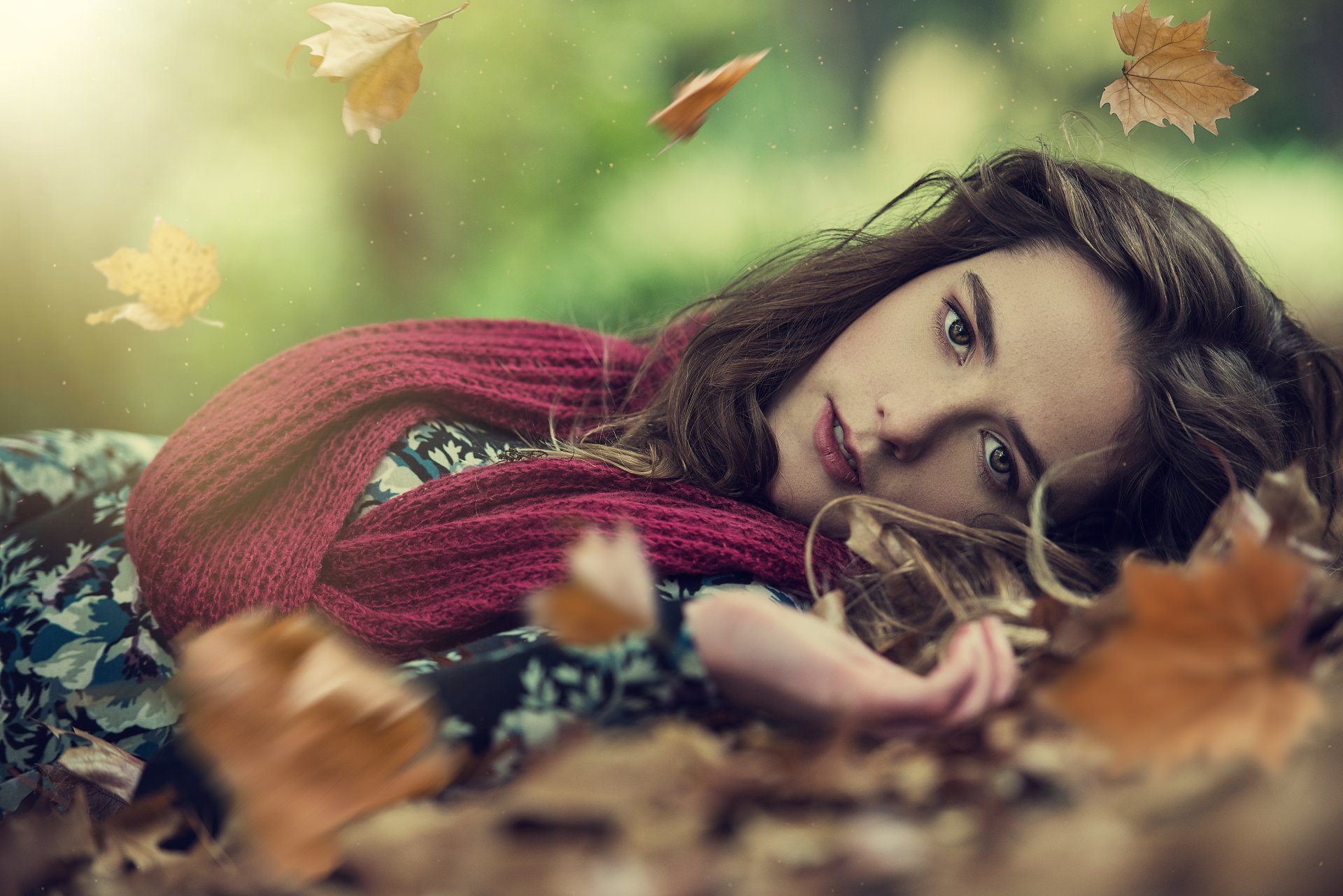 A woman in a cozy scarf lies among fallen leaves, surrounded by a serene autumn atmosphere. This HD desktop wallpaper captures a tranquil fall mood beautifully.