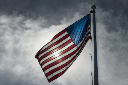 HD PC desktop wallpaper featuring a man-made American flag waving against a cloudy sky with sunlight behind it.
