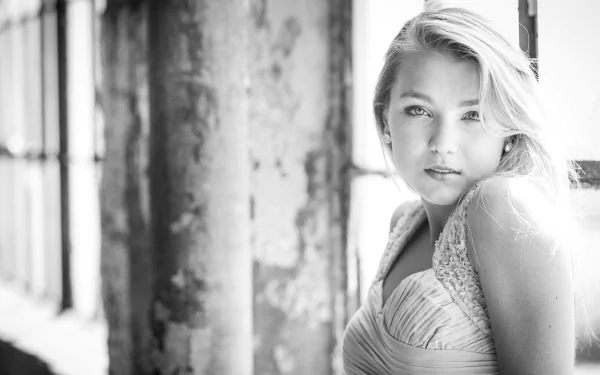 Black and white HD wallpaper featuring a beautiful woman with light hair, leaning against a rustic wall and gazing into the camera. Natural light floods the scene, highlighting her elegant features.