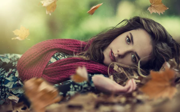A woman in a cozy scarf lies among fallen leaves, surrounded by a serene autumn atmosphere. This HD desktop wallpaper captures a tranquil fall mood beautifully.