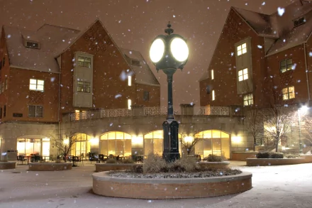 A snow-covered courtyard at night features a vintage lamp post, illuminating the surrounding modern buildings with warm light, creating a serene winter atmosphere.