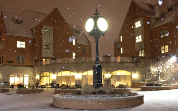 A snow-covered courtyard at night features a vintage lamp post, illuminating the surrounding modern buildings with warm light, creating a serene winter atmosphere.