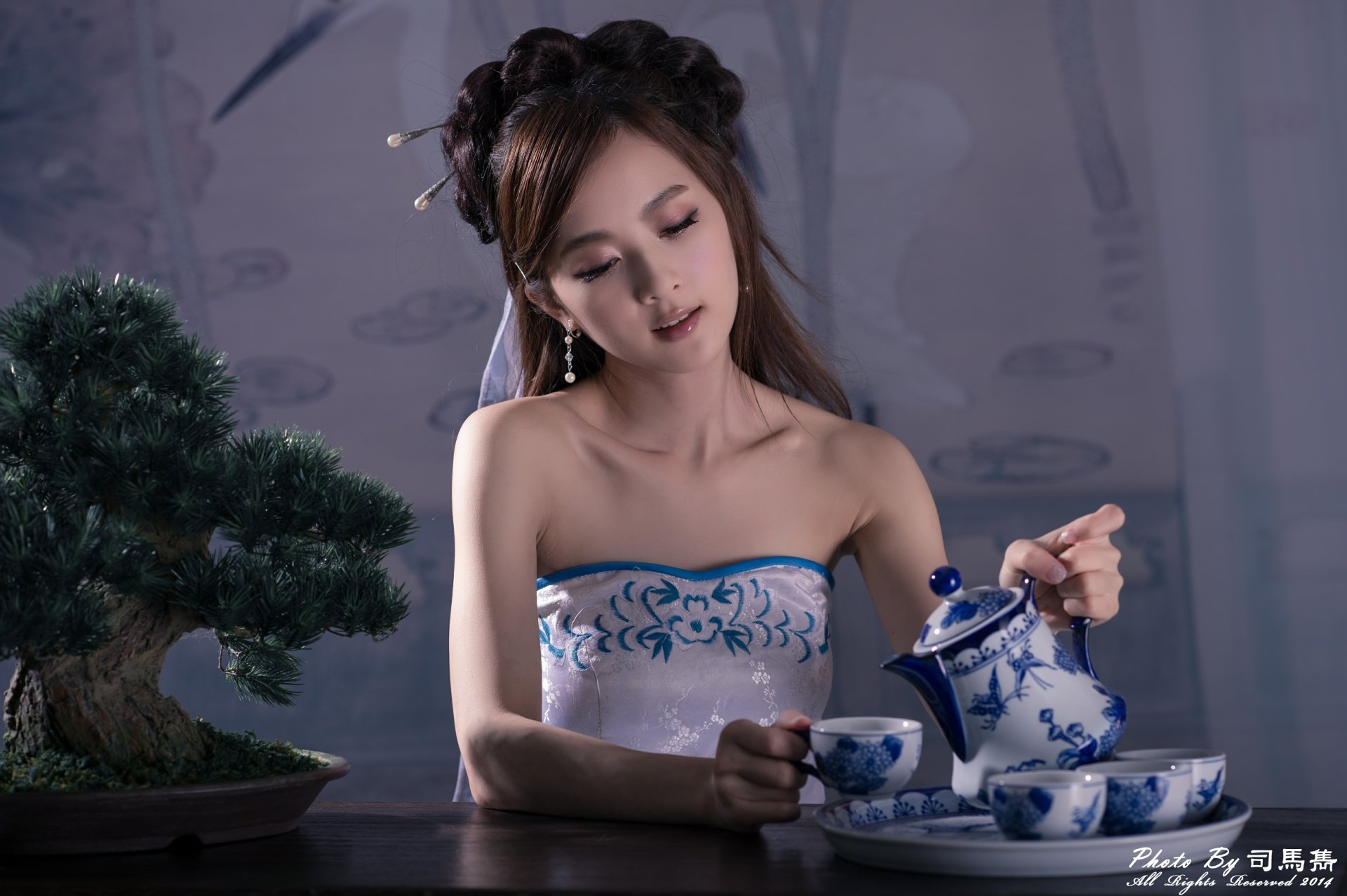 A Taiwanese woman in traditional Chinese dress and hairpin pours tea from a blue and white cup set beside a bonsai tree, captured in 4K Ultra HD.