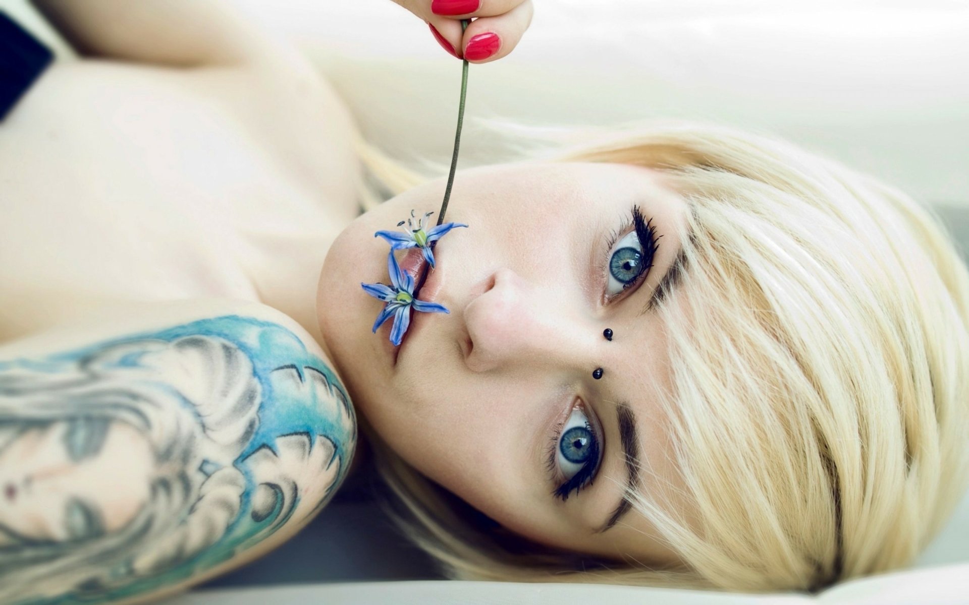 A close-up of a woman with blonde hair and captivating blue eyes, featuring a tattoo on her arm. She holds a flower delicately near her lips, creating a striking visual.