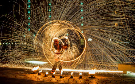 Photography of a fire-juggling performer spinning flaming poi, sparks tracing luminous circles in a long-exposure shot — 2K Quad HD PC desktop wallpaper and background.