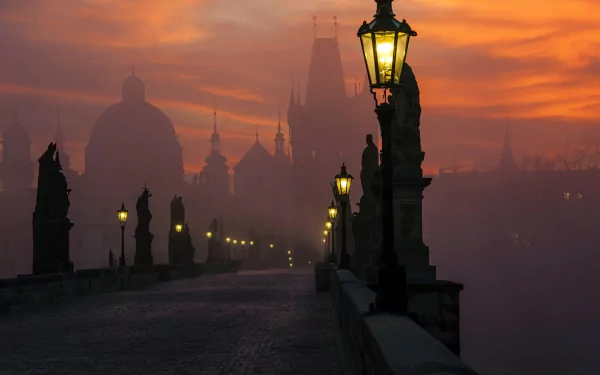 HD PC desktop wallpaper of Prague's man-made Charles Bridge at sunrise — silhouetted statues, glowing lampposts and a misty skyline.