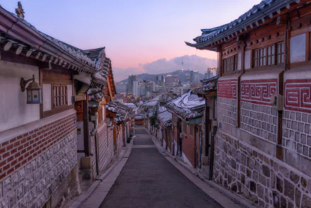 HD Desktop wallpaper of Bukchon Hanok Village in Seoul, Korea, featuring traditional Hanok houses lining a narrow street, with a view of the modern skyline in the distance under a soft pink sky.