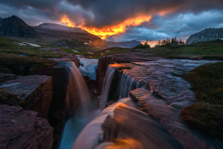 A stunning sunrise illuminates the stormy skies over a waterfall in Glacier National Park, Montana, showcasing the beauty of nature with a captivating glow.