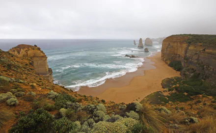 Foggy view of the Victoria coastline featuring sandy beach, ocean waves, and limestone stacks of The Twelve Apostles in Australia.