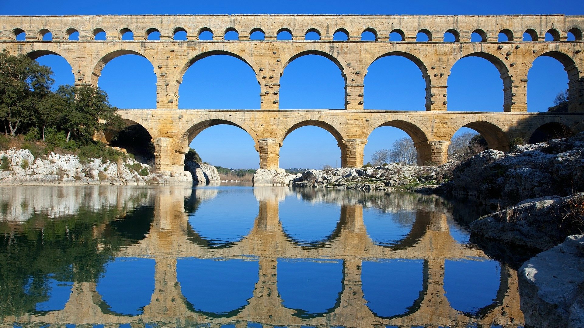 HD PC desktop wallpaper/background: stone aqueduct building over a river, calm water reflecting its arches under a vivid blue sky — architectural photography.