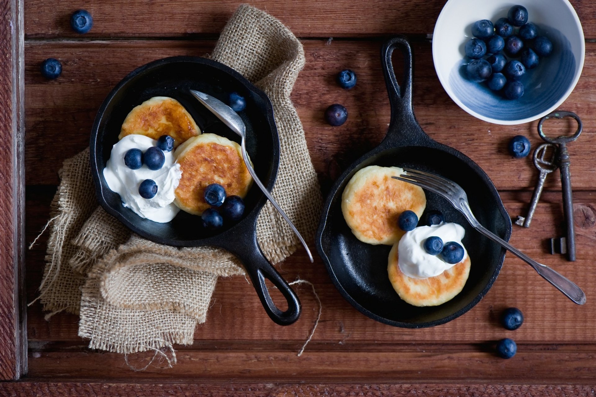 Close-up shot of blueberry pancakes topped with cream in cast iron skillets on a wooden table, captured in HD for a desktop wallpaper and background.