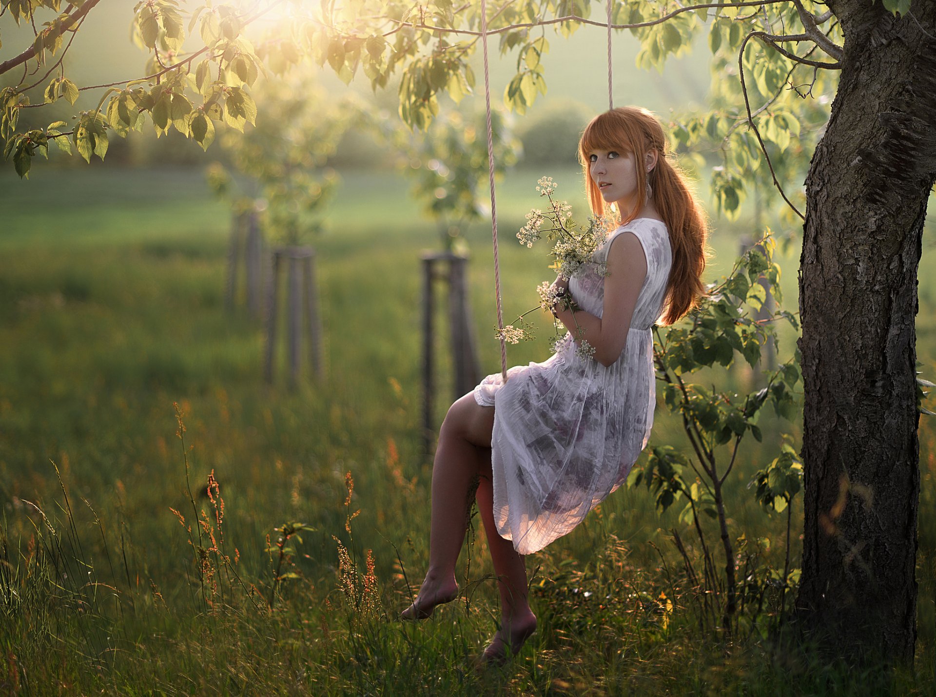 A redhead woman in a dress swings near a tree in a sunny, grassy field, holding a bouquet of wildflowers. This HD desktop wallpaper portrays a serene, nature-filled background.