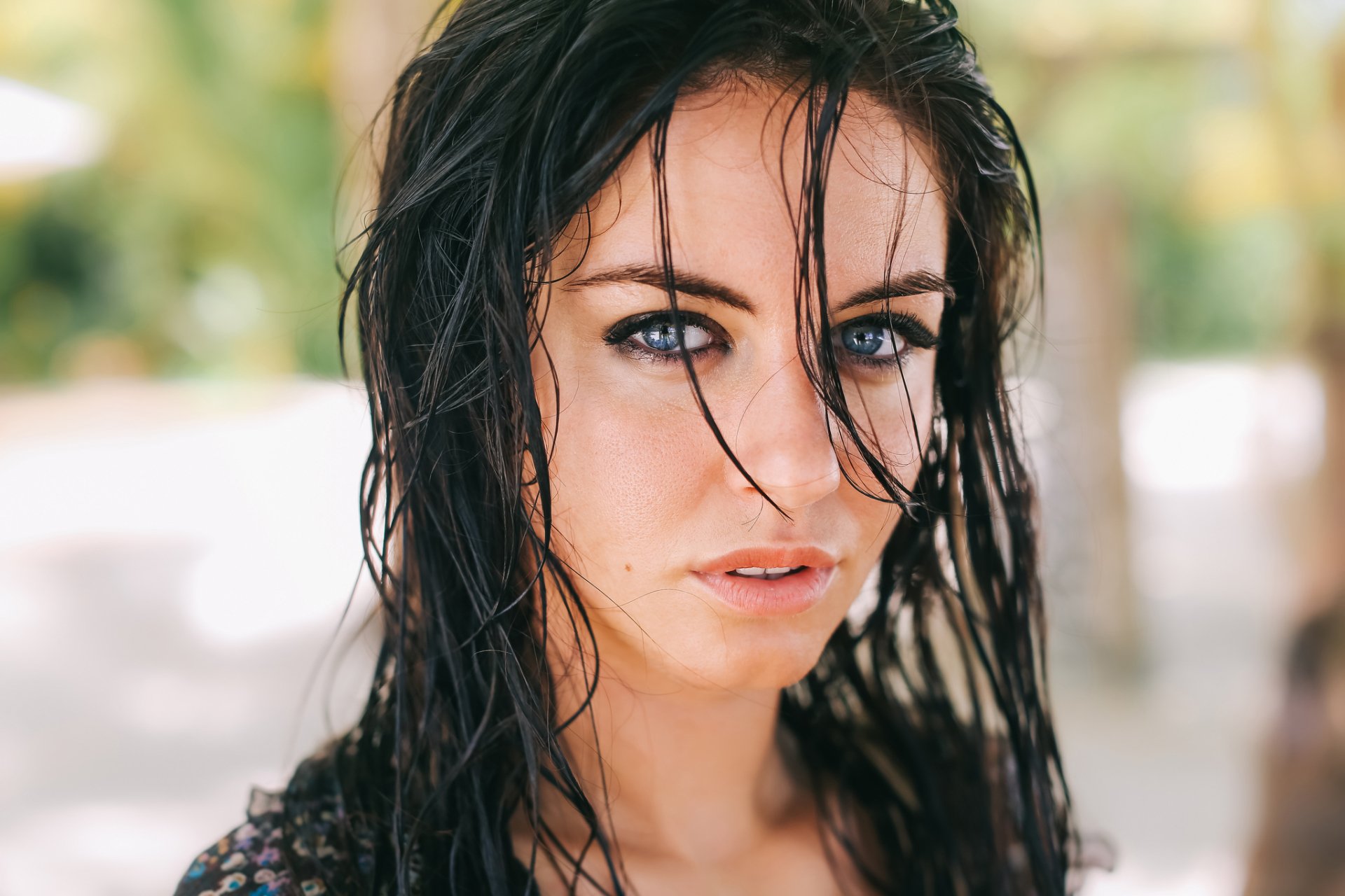 A brunette model with striking blue eyes gazes directly at the camera, her wet hair framing her face against a soft, natural background. HD desktop wallpaper.