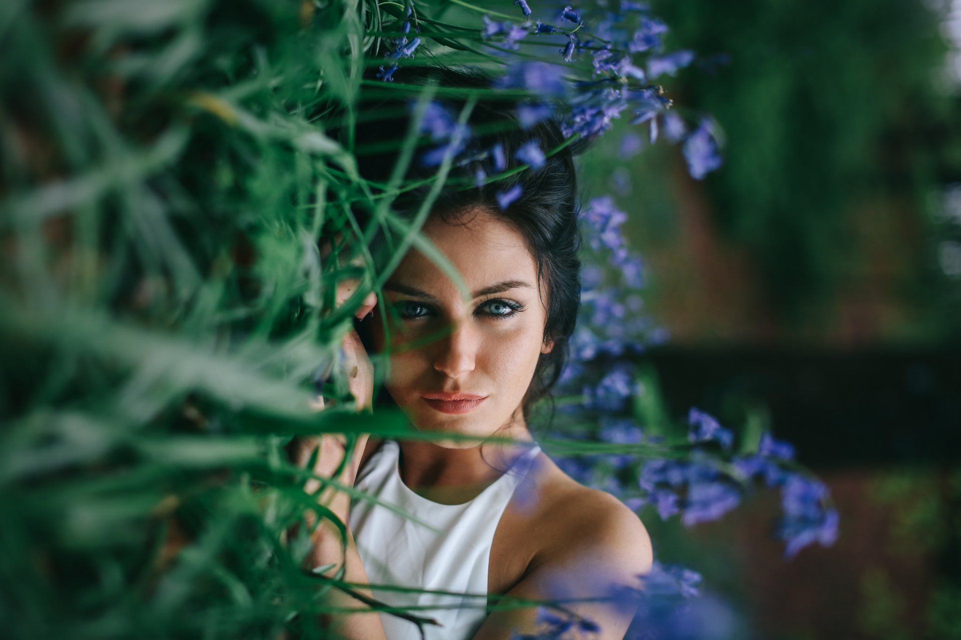 A brunette woman with blue eyes lies in the grass beside blue flowers, creating a striking HD desktop wallpaper and background.