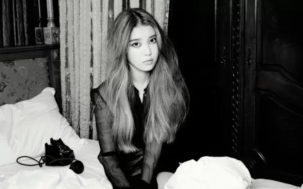 Black and white image of South Korean singer Lee Ji-eun, known as IU, sitting on a bed with a contemplative expression, showcasing her distinctive brunette hair and artistic vibe.