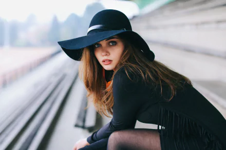 HD desktop wallpaper featuring a brunette model with blue eyes, wearing a black outfit and wide-brimmed hat, sitting on outdoor steps. The background is softly blurred, focusing on her intense gaze.