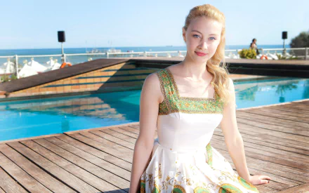 HD wallpaper of Canadian actress Sarah Gadon with blonde hair and blue eyes, posing outdoors by a pool under clear skies.