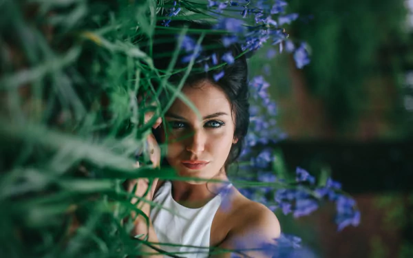 A brunette woman with blue eyes lies in the grass beside blue flowers, creating a striking HD desktop wallpaper and background.