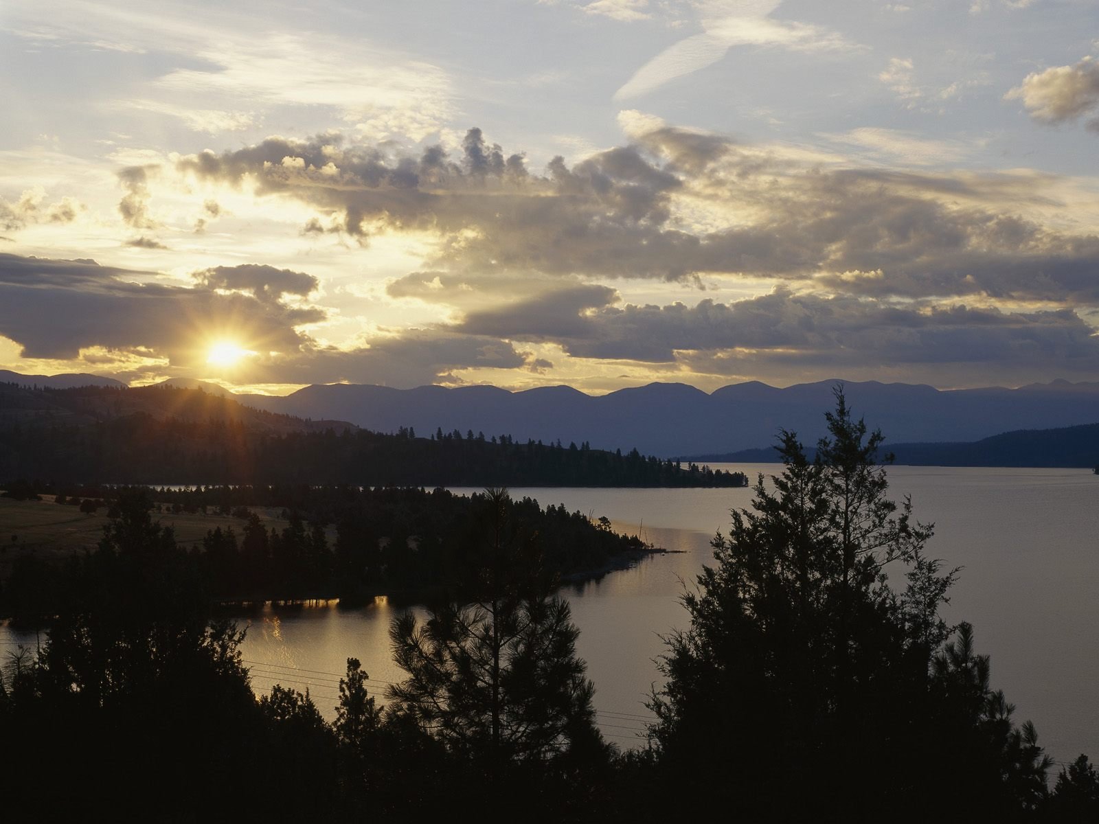 HD nature desktop wallpaper showing a serene lake at sunset with silhouetted trees and mountains under a partly cloudy sky.