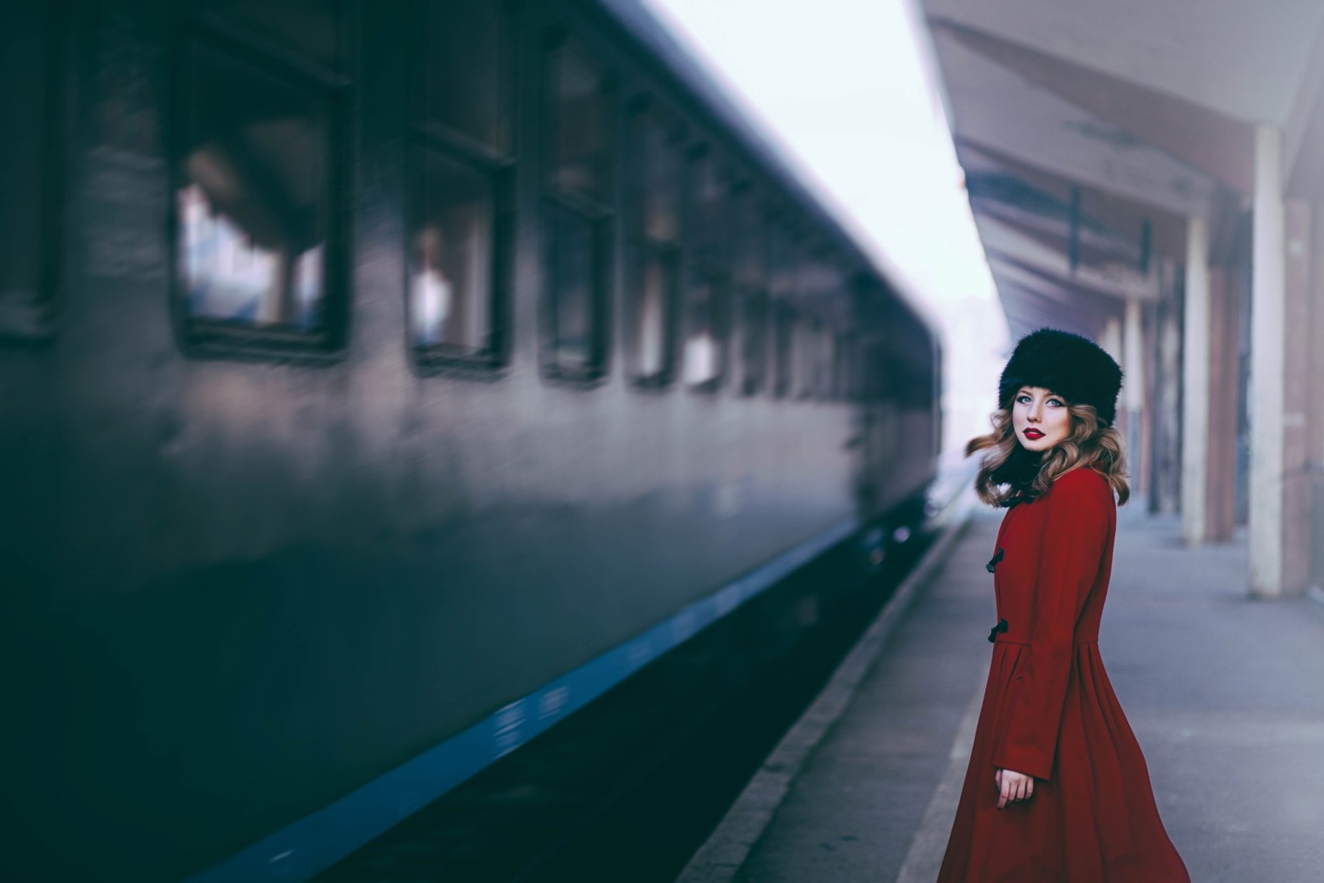 A woman model wearing a fur hat and red coat stands on a train station platform beside a dark blue train, captured in a high-definition desktop wallpaper.