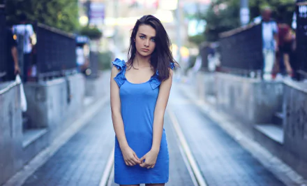 A brunette woman with black hair and blue eyes, wearing a blue dress, stands in a city setting with a blurred background. This HD image features a depth of field effect and a central model.
