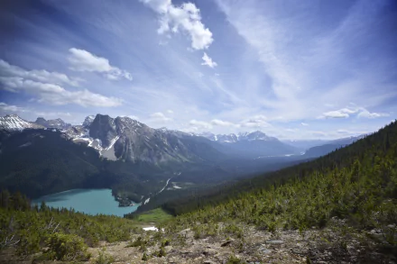 A breathtaking landscape of British Columbia's Canadian Rockies, featuring a serene lake framed by lush greenery and towering mountains under a blue sky with wispy clouds.