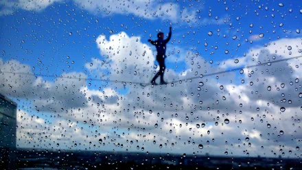 A tightrope walker balances against a backdrop of blue skies and fluffy clouds, viewed through a rain-speckled window, creating a dramatic and inspiring photography scene.