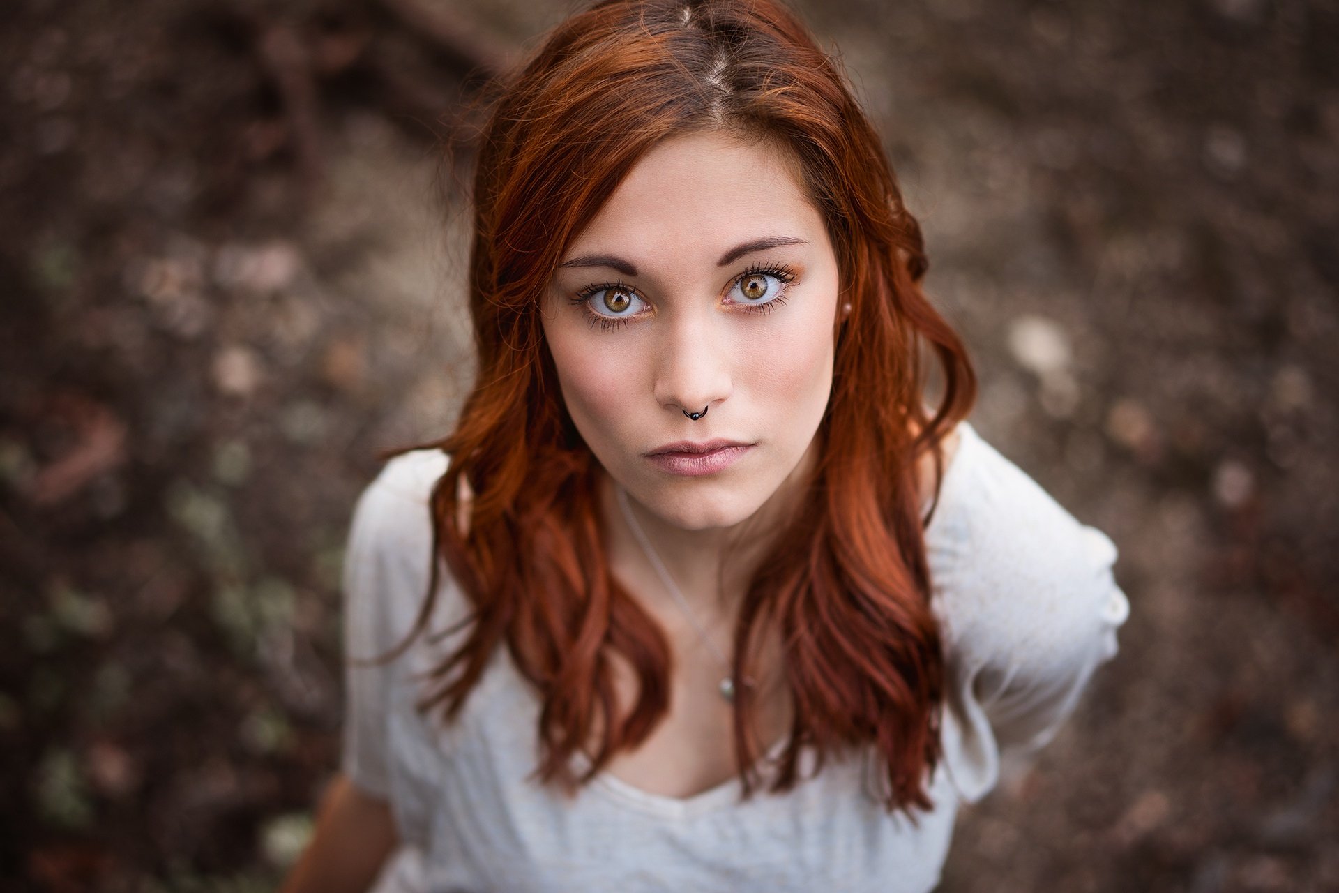 A striking model with long red hair and brown eyes gazes directly at the camera, surrounded by a soft bokeh background, creating an engaging HD wallpaper for desktop use.
