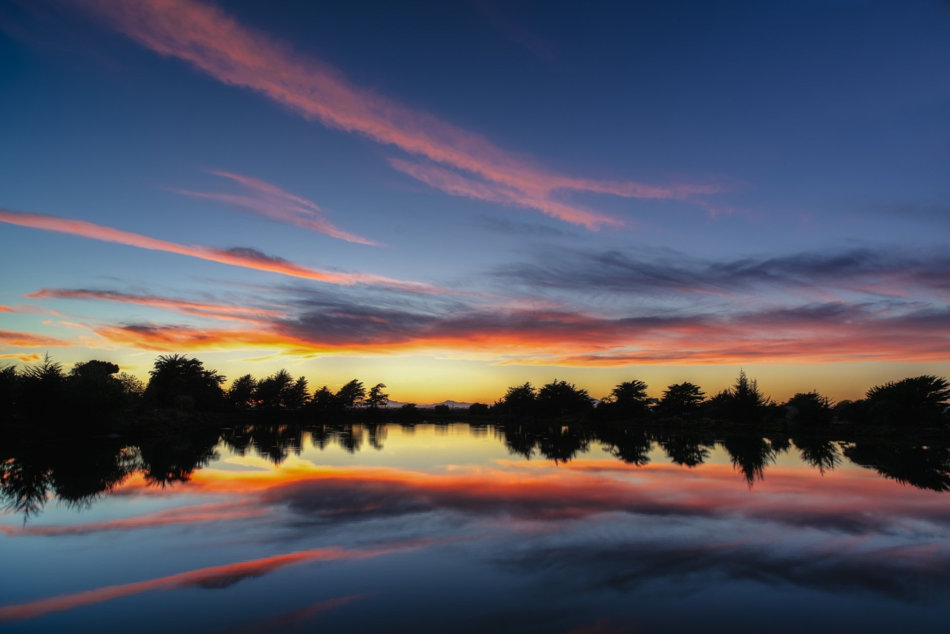 HD desktop wallpaper capturing a tranquil sunset over a landscape with silhouetted trees, clouds, and vibrant reflections on the calm water surface.